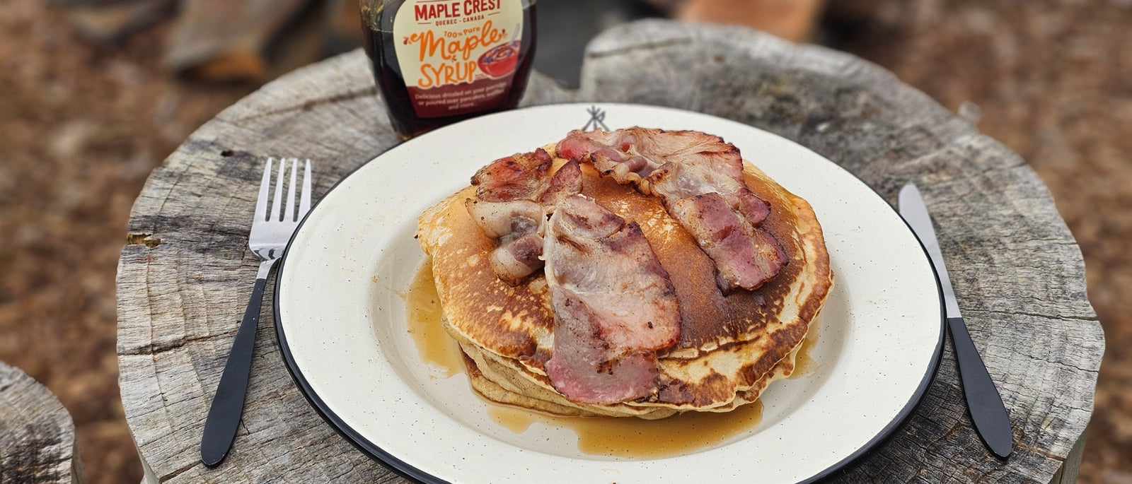 Stack of American pancakes topped with grilled bacon and maple syrup on an enamel camp plate, served on a log stump outdoors