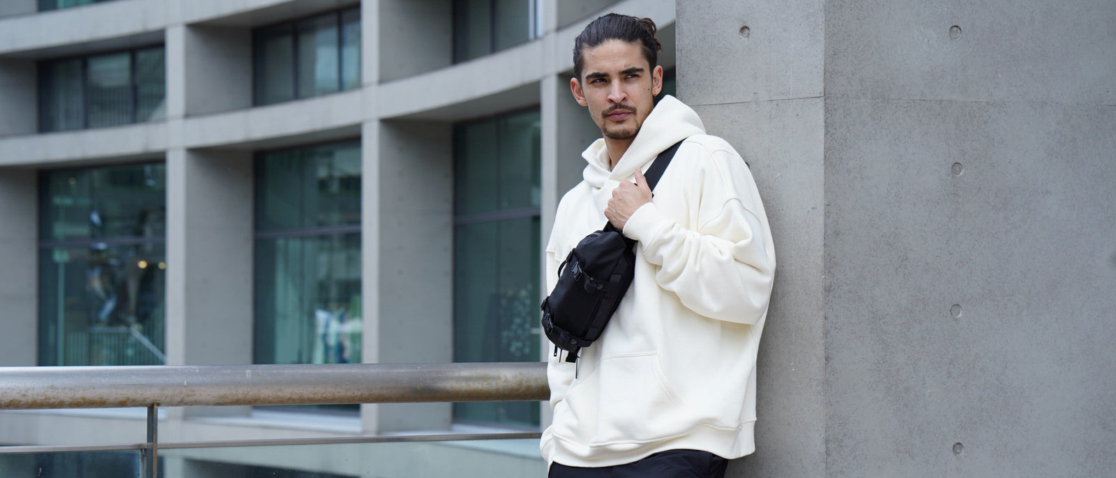 A man wearing a cream oversized hoodie and black trousers leans against a concrete pillar in a modern city setting, wearing a black CODE OF BELL sling crossbody.