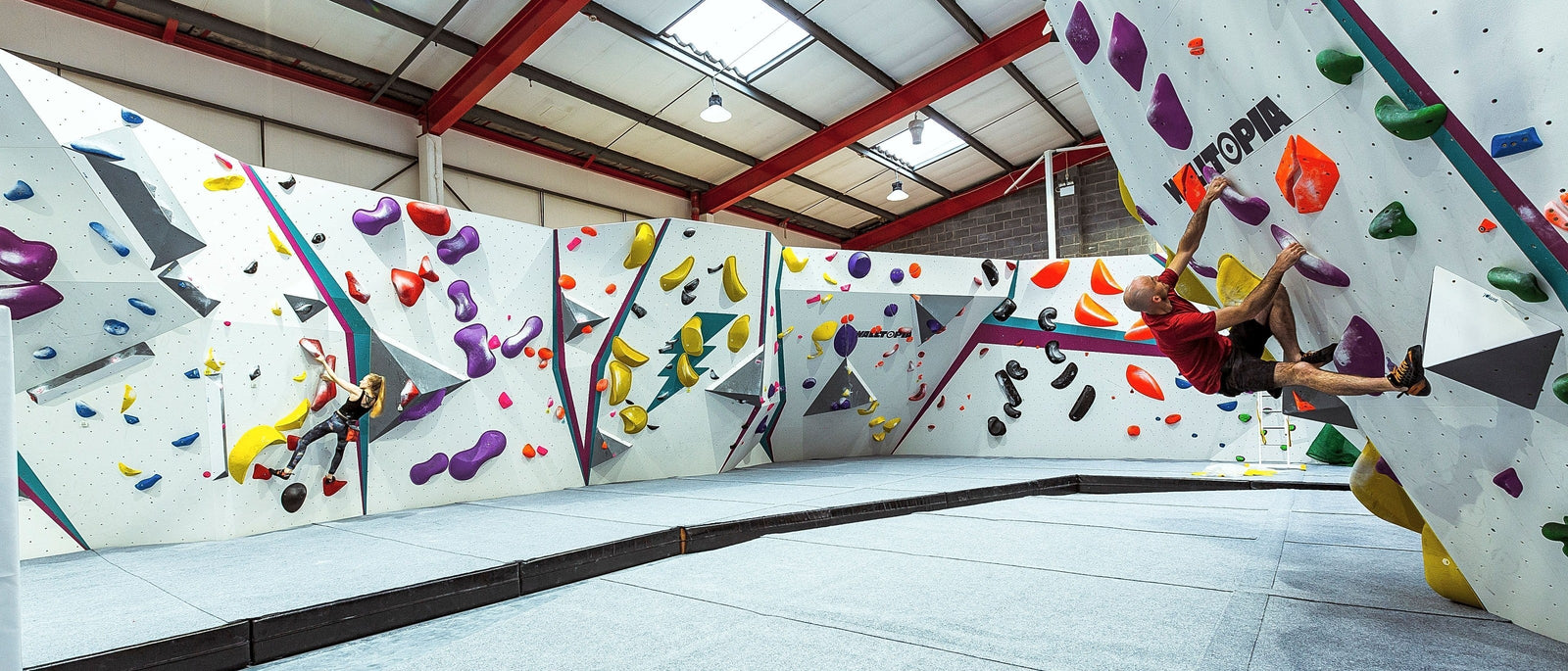 A climber dynos on an overhanging bouldering wall at Flash Point climbing centre in Bristol, with a second climber working a vertical problem in the background amid colourful holds and geometric wall art.