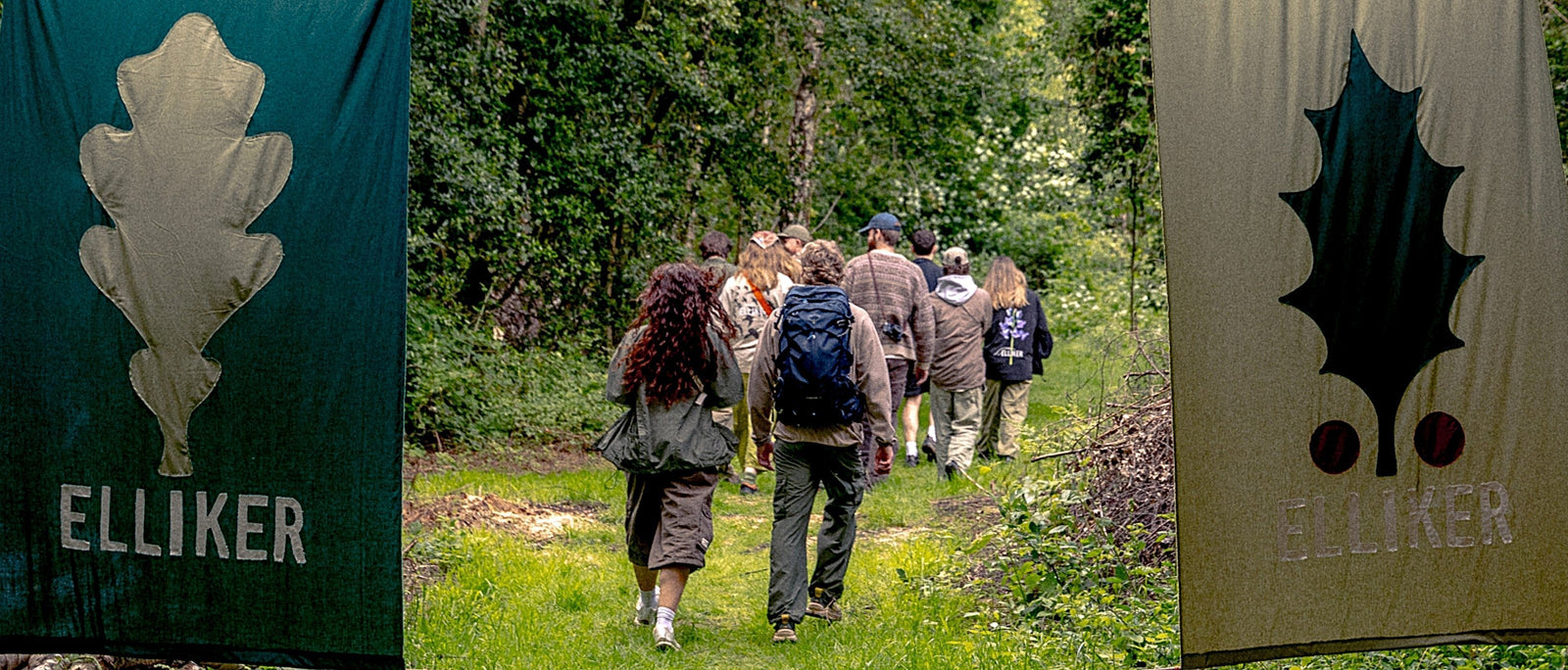 A group of people walk through a gap between two large fabric banners printed with leaf motifs and the Elliker logo, heading into dense green woodland on a summer's day.