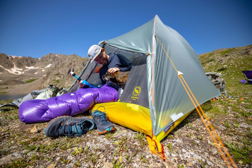 Hiker inside a grey and yellow tent with a purple sleeping bag and backpack outside on an alpine meadow.