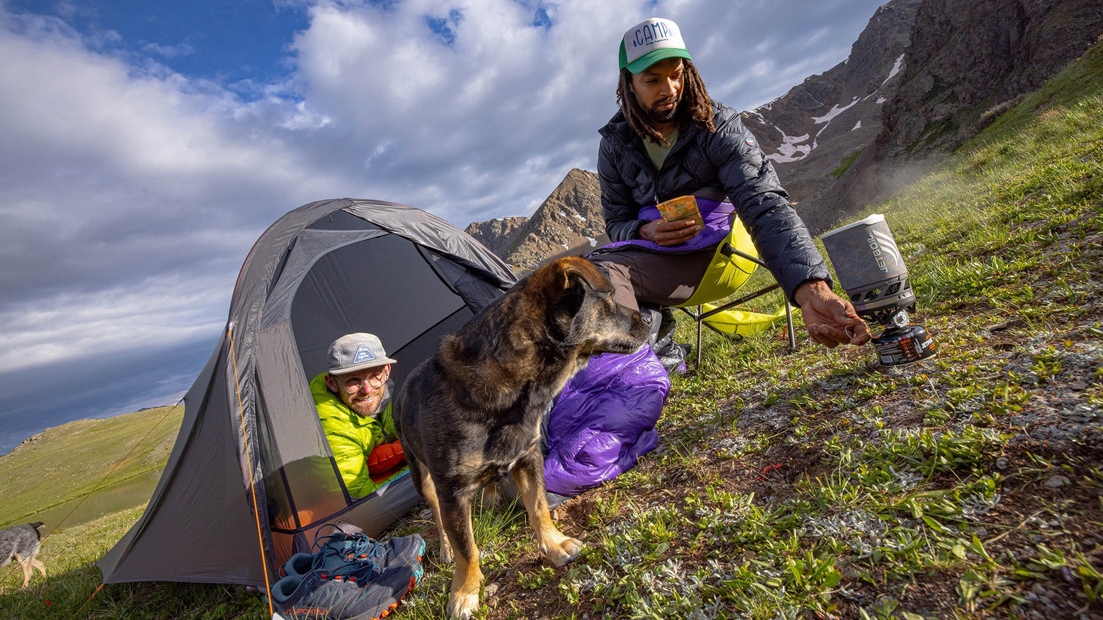 Two campers and a dog at a tent on a mountainside with dramatic cloudy skies and rocky peaks in the background.