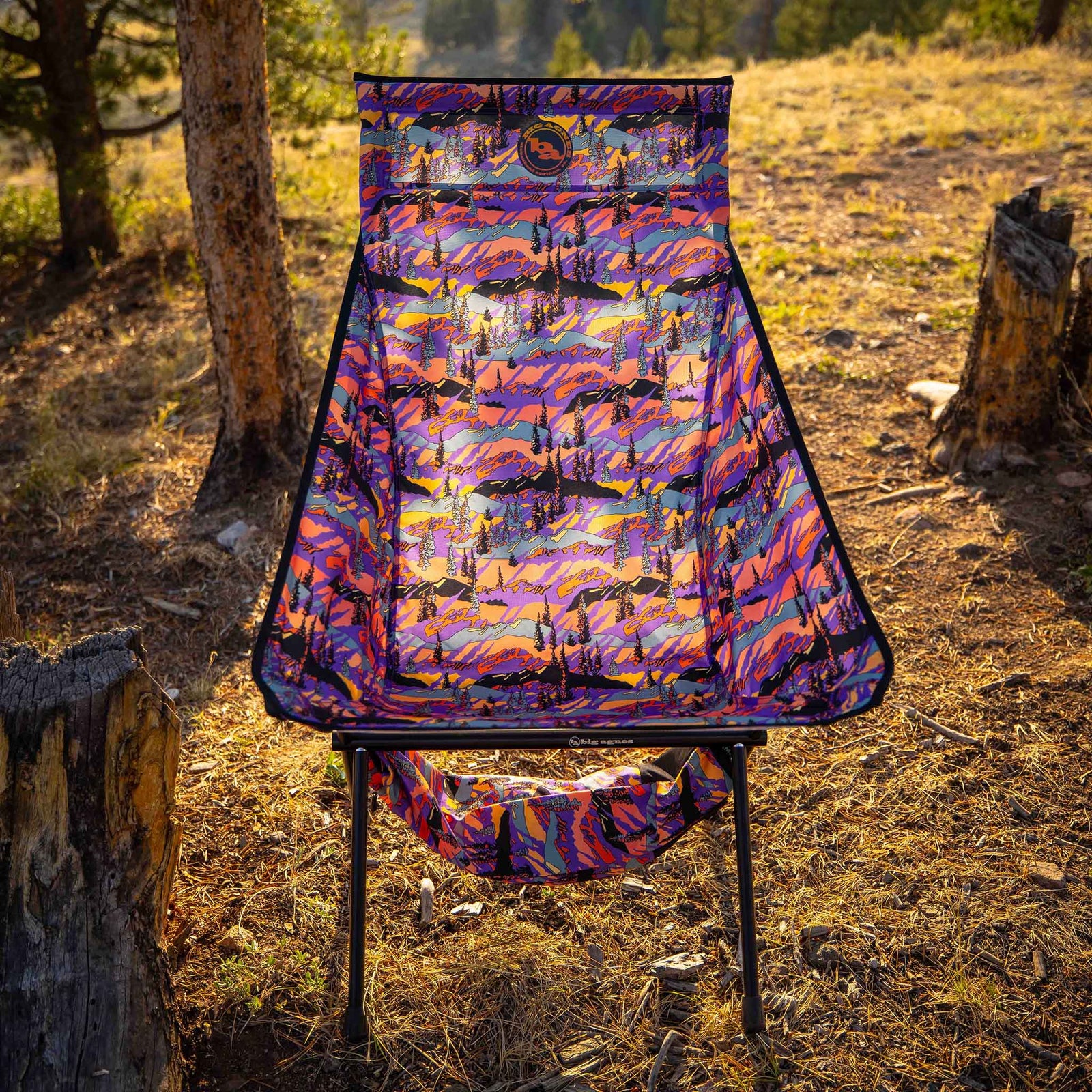Helinox camping chair with vibrant purple and orange sunset landscape print, set up in a forest clearing.