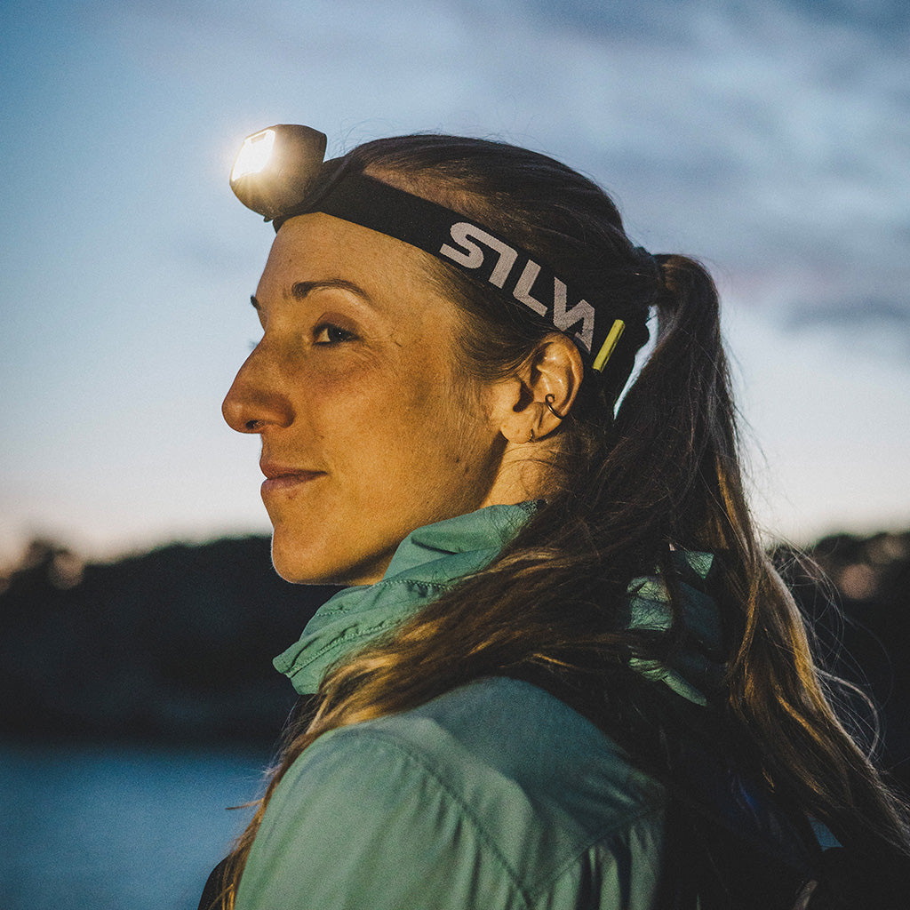 Person wearing a head torch headband by a lake at dusk, looking outdoors.