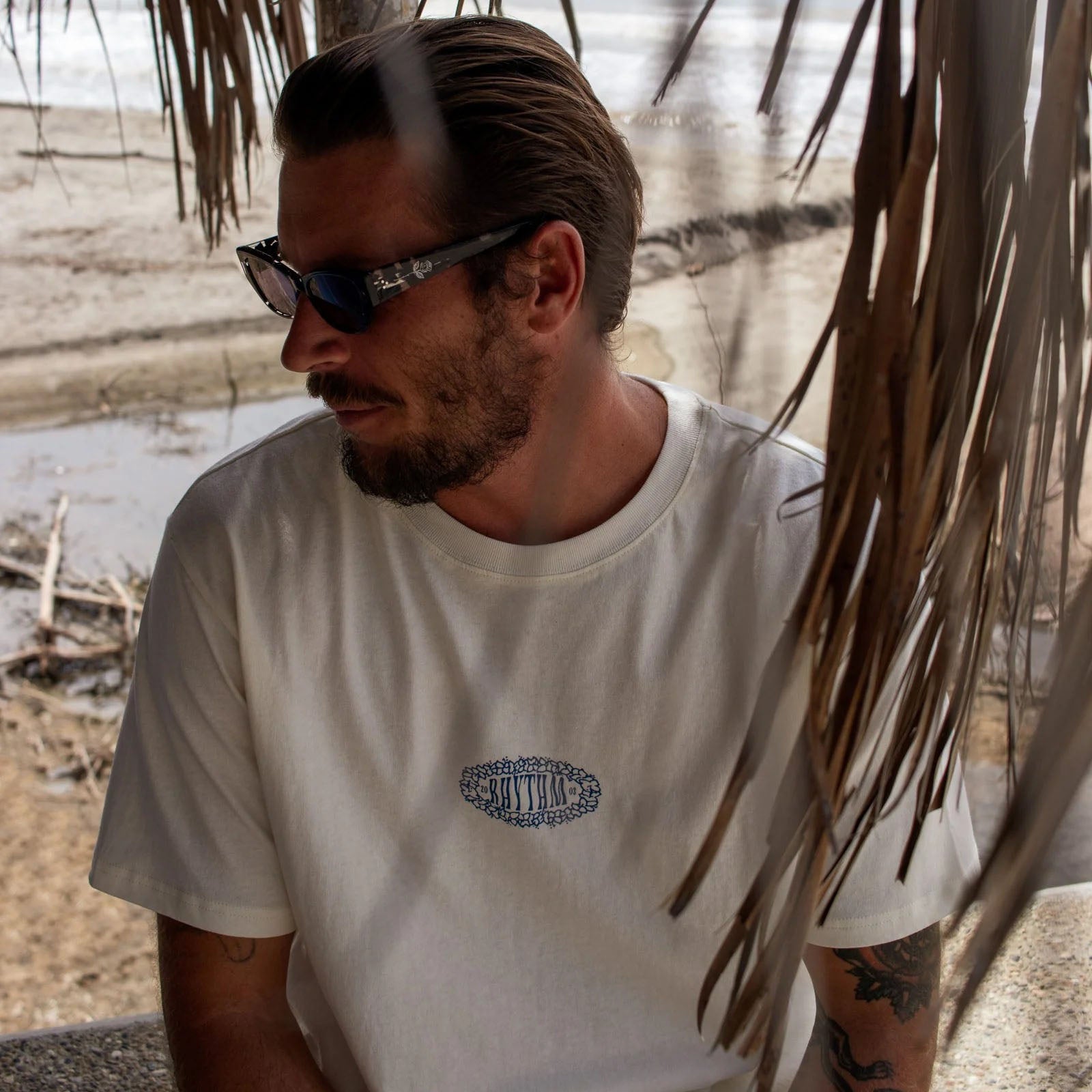 Man wearing cream t-shirt and sunglasses under palm fronds on a beach.