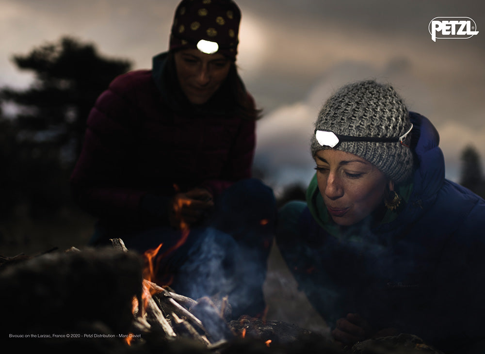 Two people wearing Petzl headlamps and winter hats tend a campfire at dusk in the Larzac, France.
