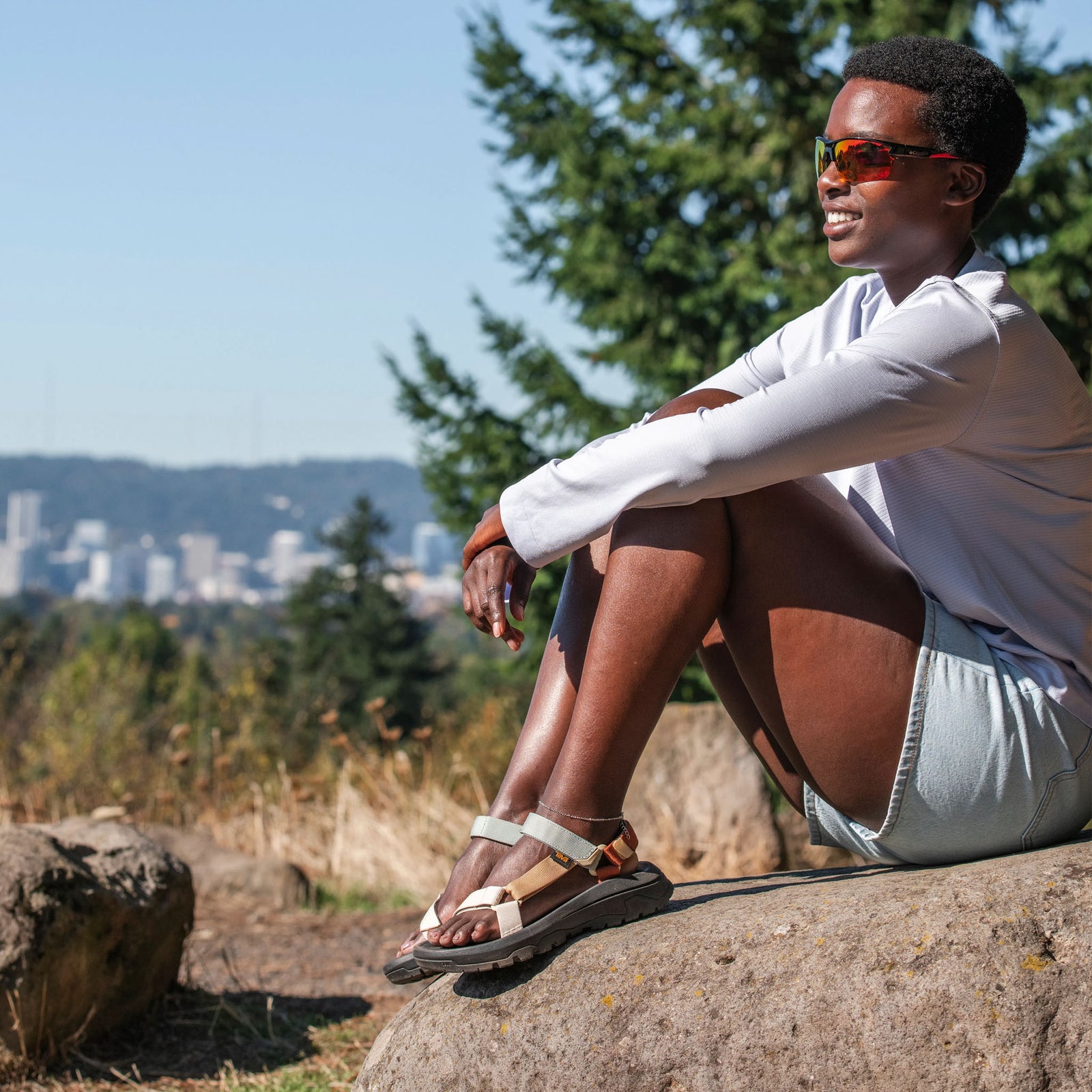 Person wearing Teva sandals and white long-sleeved top sitting on a rock overlooking a forested valley