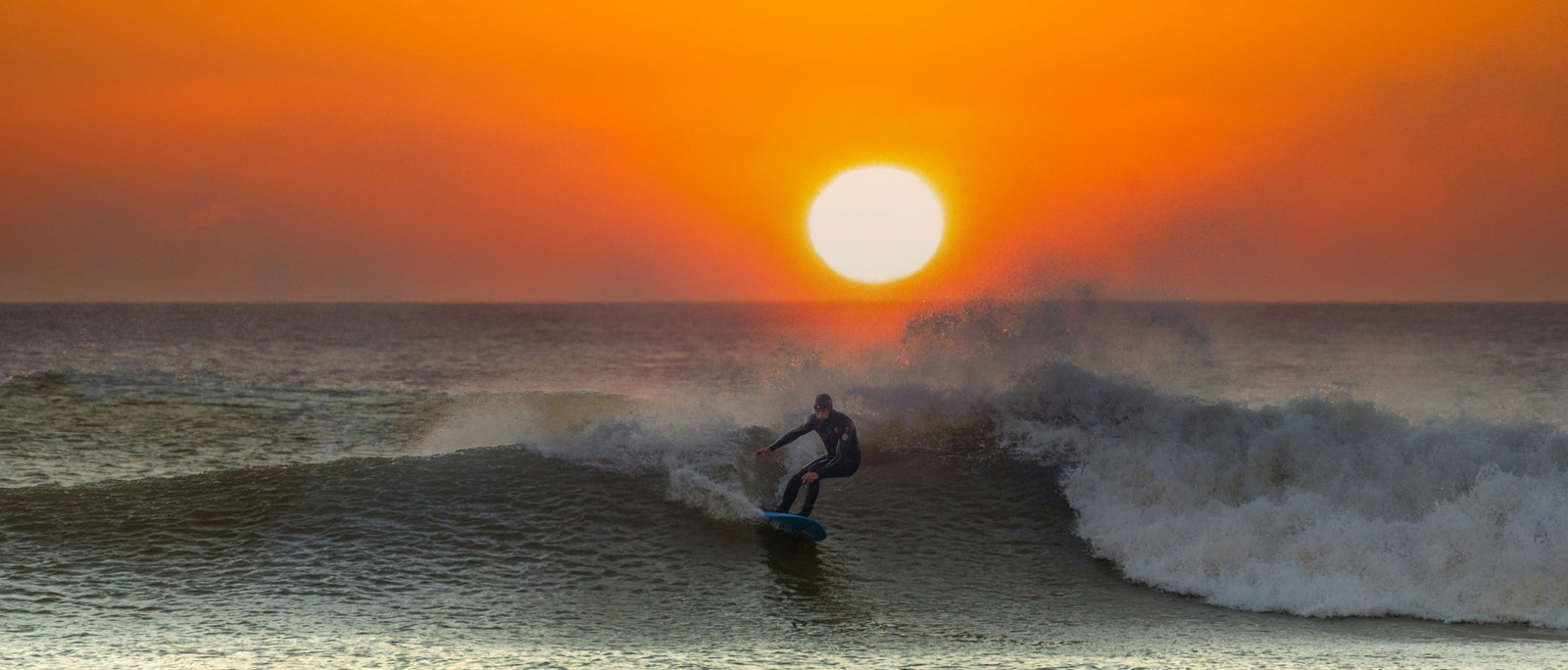 Surfer riding a wave beneath a vivid orange sunset, Southerndown Beach (Dunraven Bay), Vale of Glamorgan, Wales