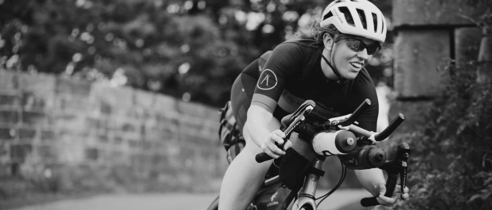 black and white photo of a woman cycling round a corner near a low stone wall