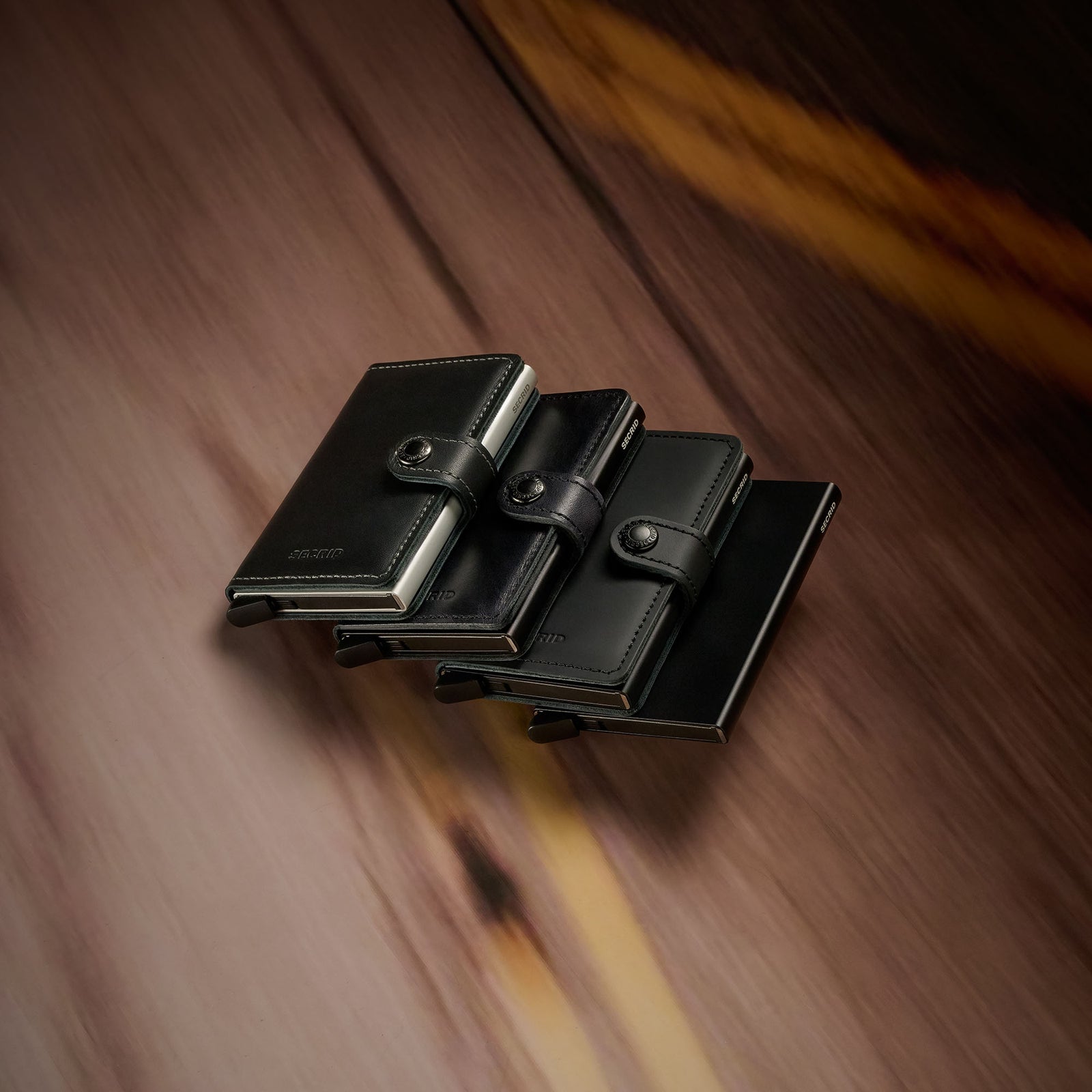 Four black leather wallets with press-stud closures arranged in a cascading display on a neutral fabric background.
