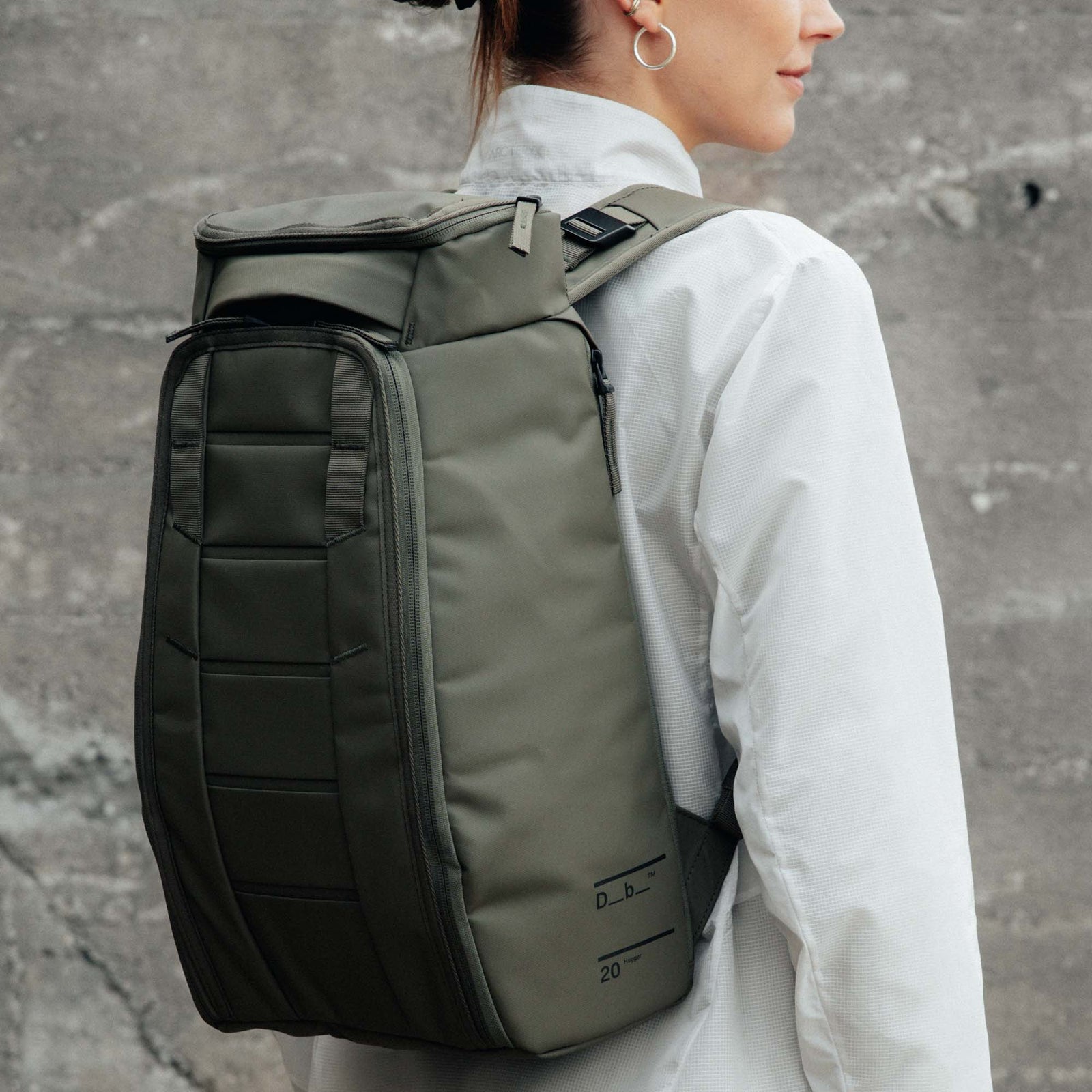 Woman wearing olive green and black backpack with roll-top closure against concrete wall.