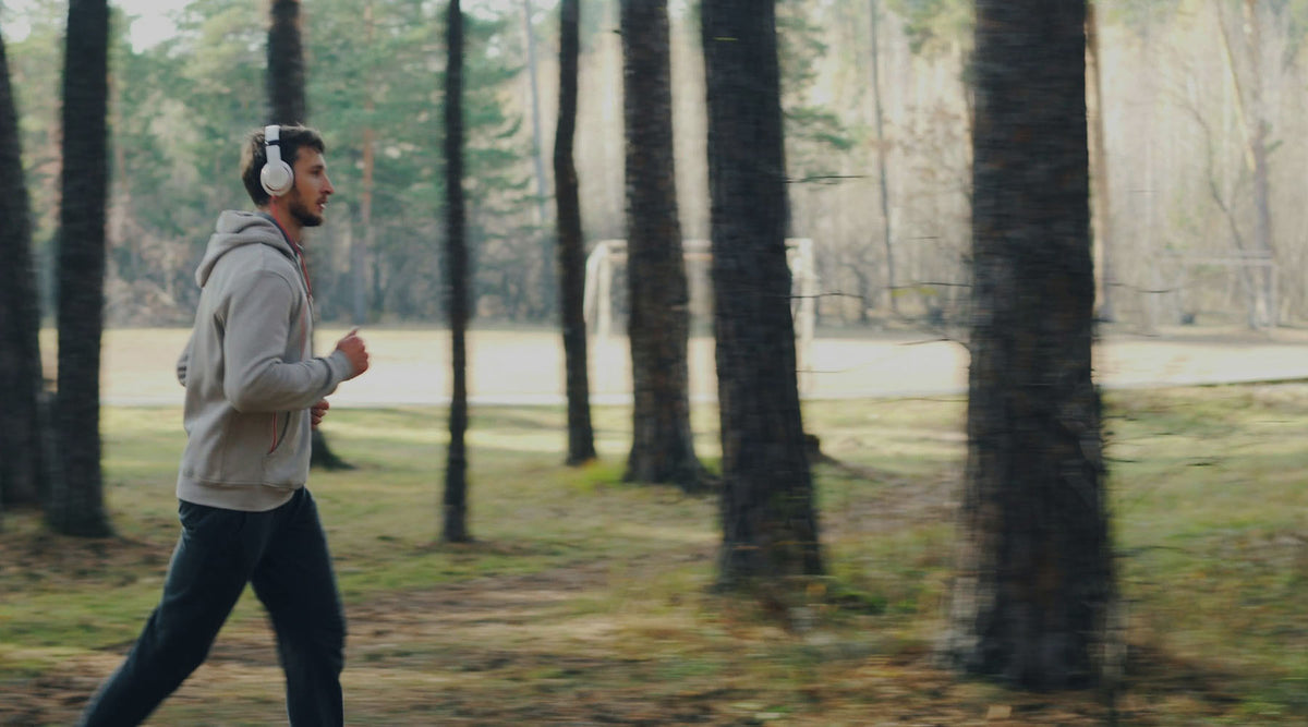 A man jogging through a pine forest while wearing headphones and a grey hoodie.