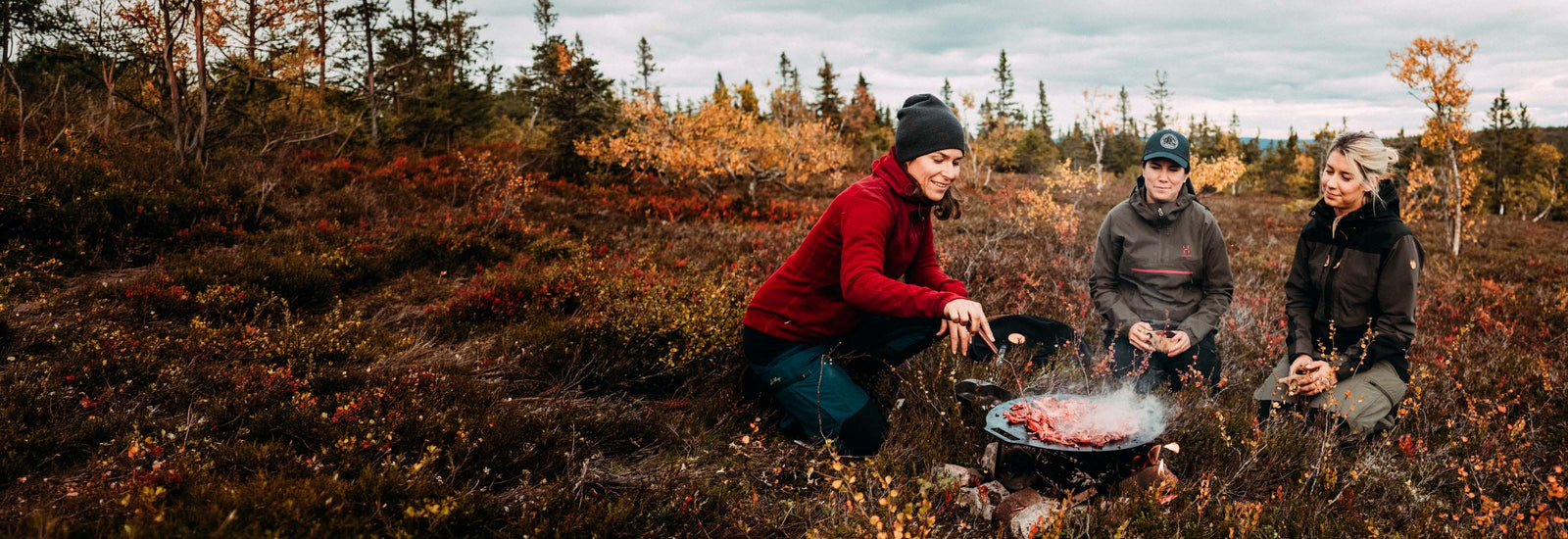 Three people gathered around a campfire in autumn woodland, cooking outdoors in fall landscape.