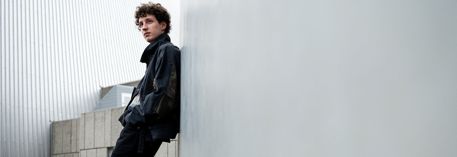 Person wearing a black Deus Ex Machina jacket against a corrugated metal wall.