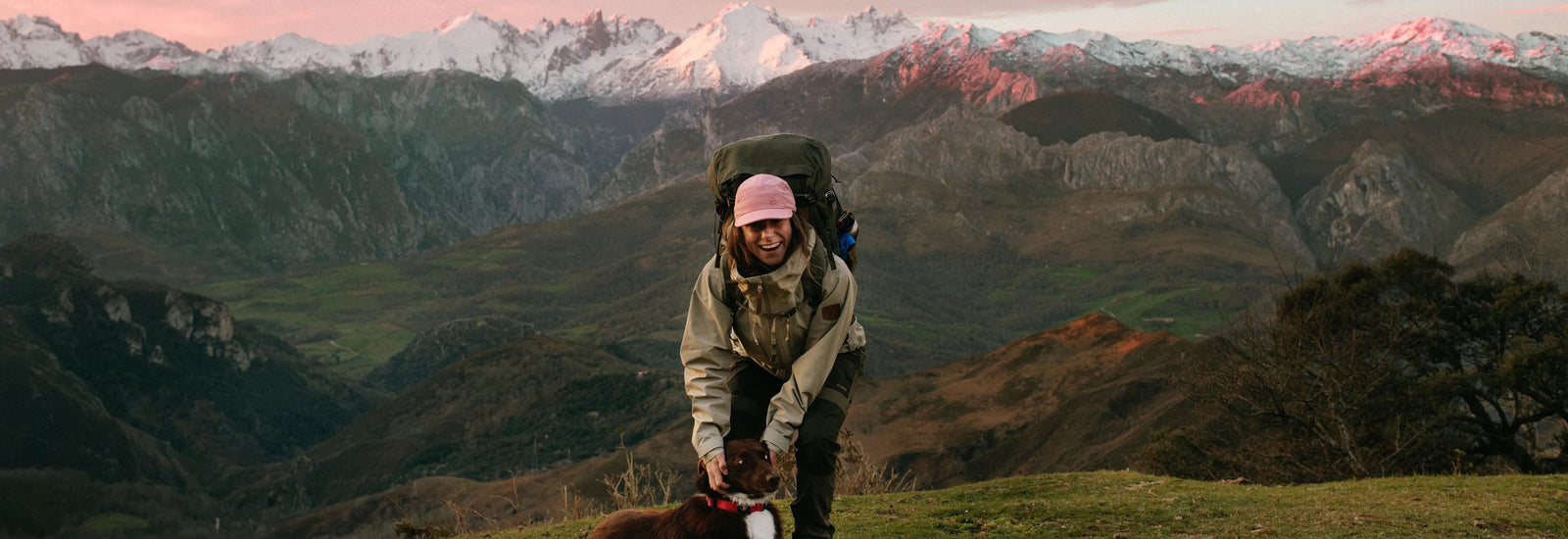 A hiker wearing a Fjällraven backpack and pink beanie on a hillside overlooking a snow-capped mountain valley at sunset.