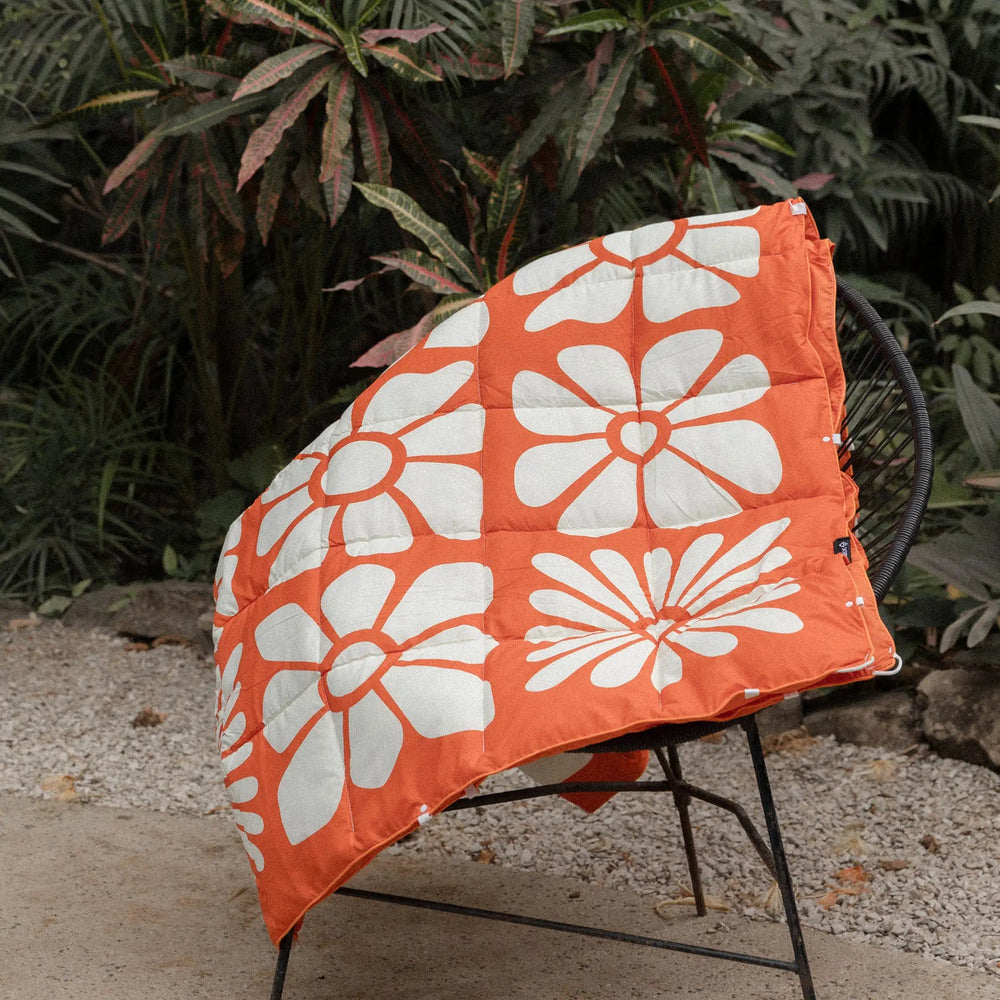 Nomadix Graphene Puffer Blanket in Big Flowers Expedition Orange draped over a camping chair against tropical foliage.