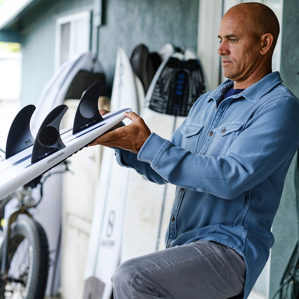 Man examining a surfboard with fins in a garage, Outerknown brand products.