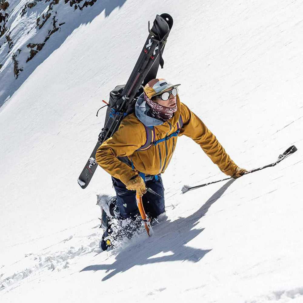 Skier in mustard jacket carrying skis down snowy mountain slope.