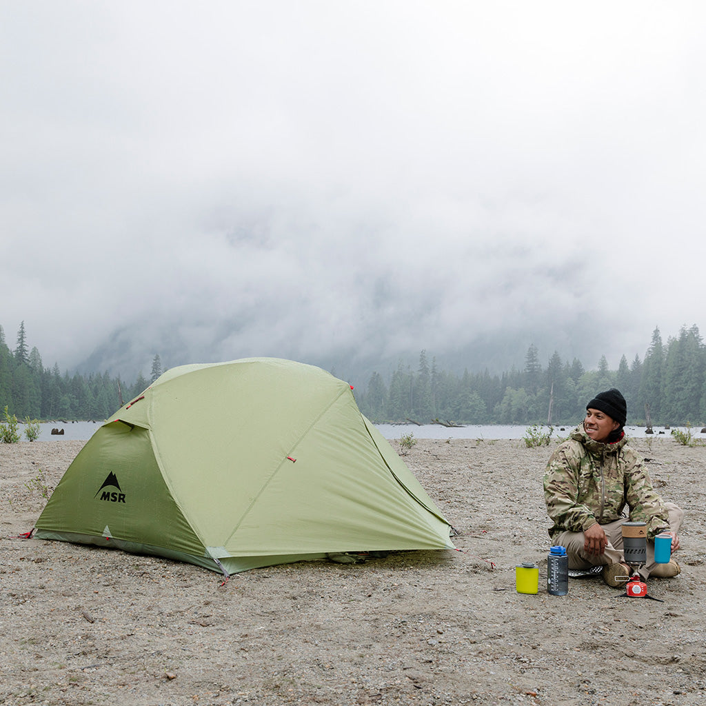 Camper with MSR tent and camping gear on misty lakeside beach, sleep sale promotion.