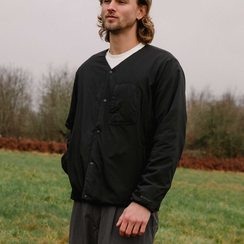 Man wearing Snow Peak water-repellent knit cardigan in black over white t-shirt, standing in rural landscape.