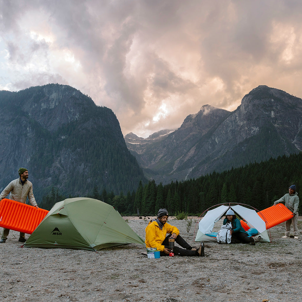 Campers with MSR tents and orange sleeping pads at a mountain valley campsite during summer.