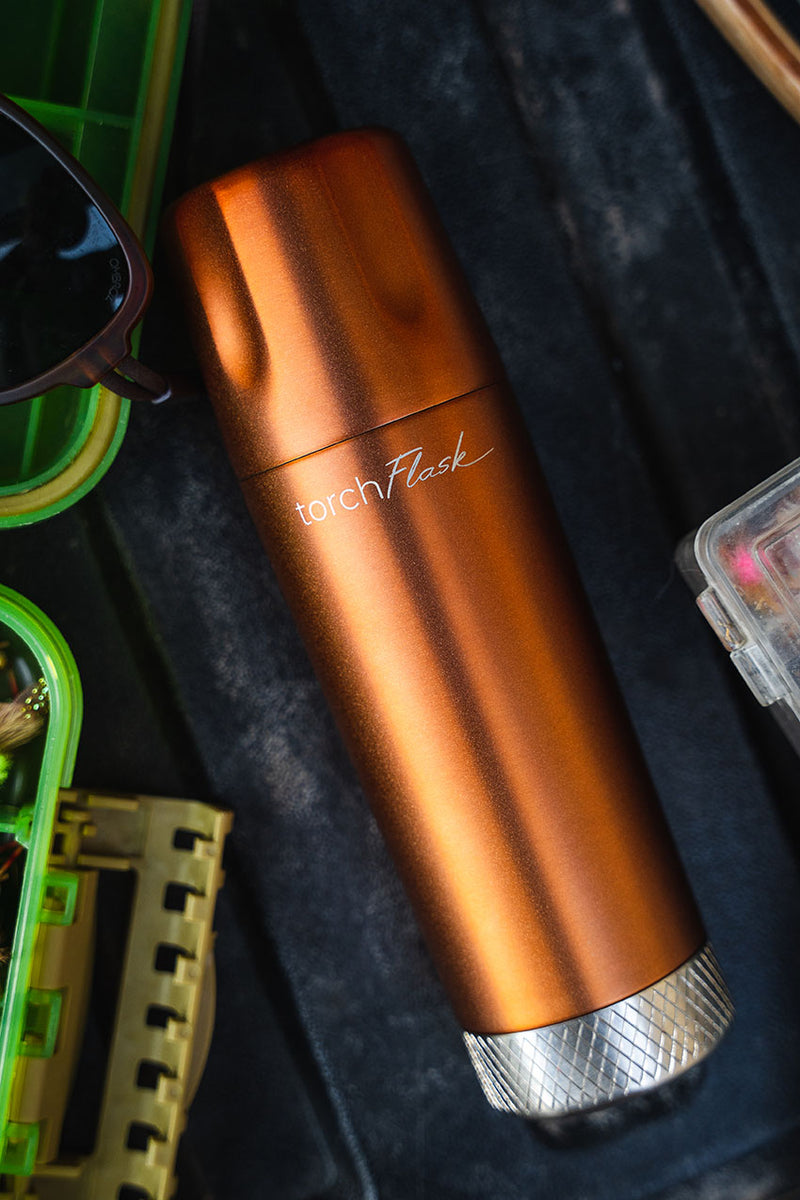 Copper torchflask bottle surrounded by outdoor gear including sunglasses and a tape measure.